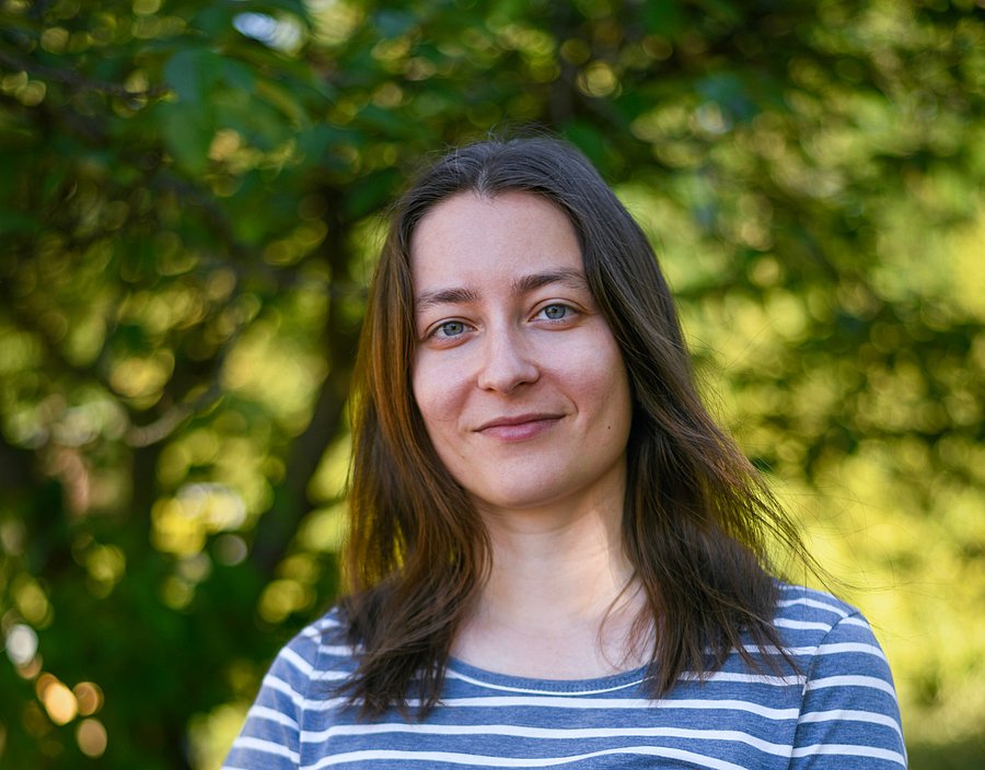 A photo of Anna Sauer. It shows a smiling young woman wearing a blue and white striped shirt, standing outdoors in front of a tree.