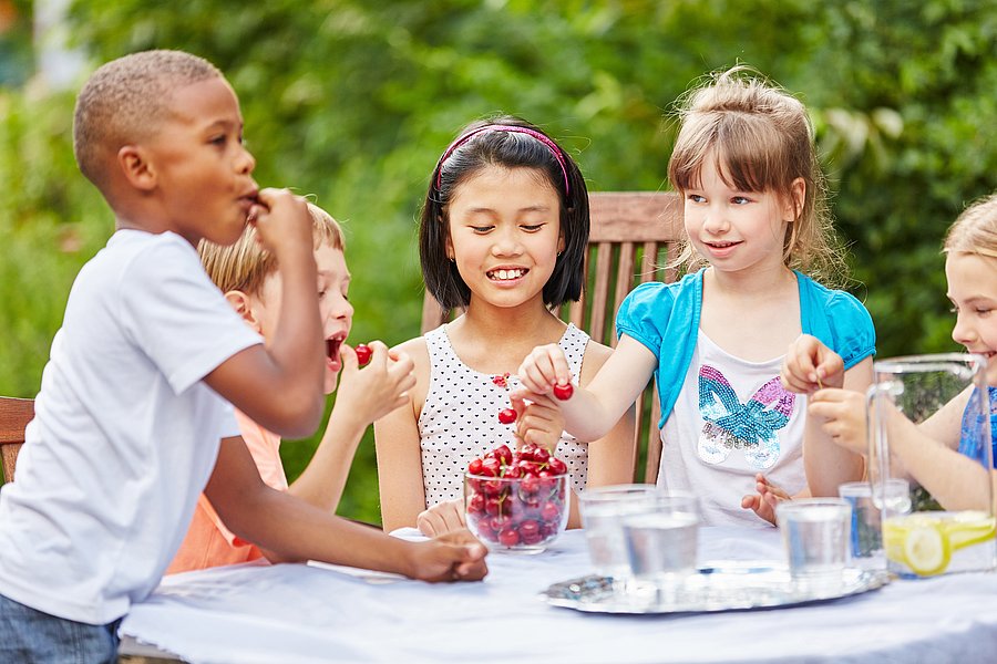 Das Foto zeigt Kinder im Garten. Sie sitzen lachend am Tisch und essen Kirschen. 