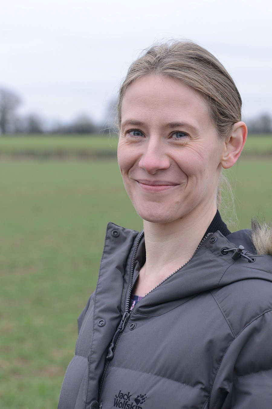 A photo of Lizzie Sagoo. It shows a woman wearing a grey jacket, standing outdoors in a grassy field on a cloudy day.