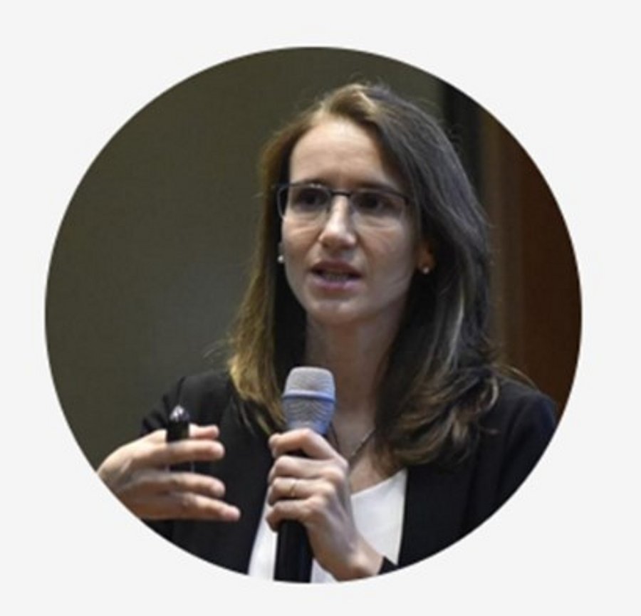 A photo of Virgina Pravia. It shows a woman with brown hair giving a presentation. She wears a black jacket over a whte shirt and glasses. She holds a microphone in her left hand and a remote control in her right hand. A presentation screen is visible in the background.