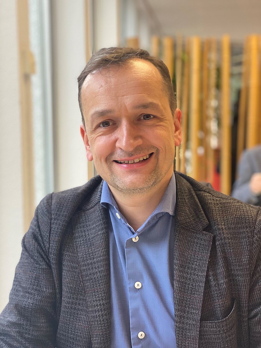 A photo of Christian Holzleitner. It shows a middle-aged man smiling into the camera. He has brown hair and blue eyes. He wears a suit jacket over a blue shirt.
