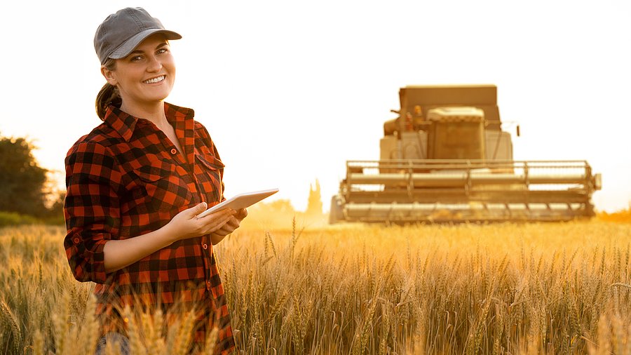 Eine Landwirtin steht in einem Getreidefeld. Sie trägt ein Tablet in der Hand und schaut in die Kamera. Im Hintergrund fährt ein Mährdrescher. 