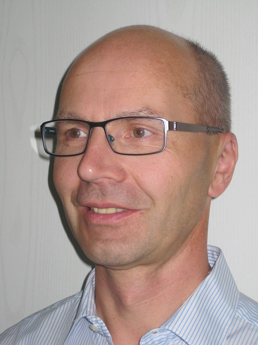 A photo of Hans Fink. It shows a smiling, balding, middle-aged man with green eyes and brown, graying hair. He wears glasses and a white shirt with blue stripes.