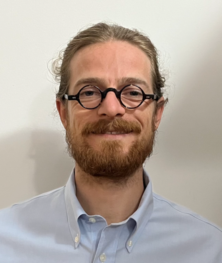 A photo of Simon Krämer. It shows a smiling Man with blonde hair, a full beard and blue eyes. He wears glasses and a light blue shirt. The background of the photo is white.