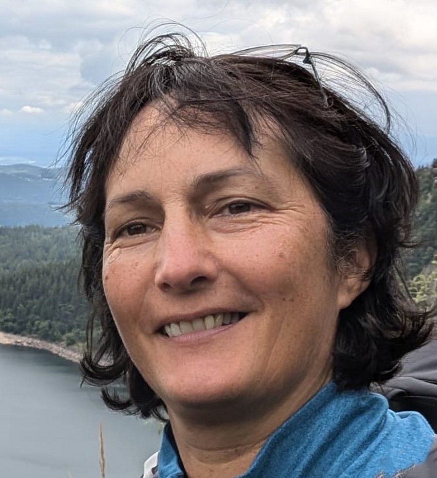 This photo shows a woman with brown hair smiling at the camera. The background of the photo is a rocky coast.