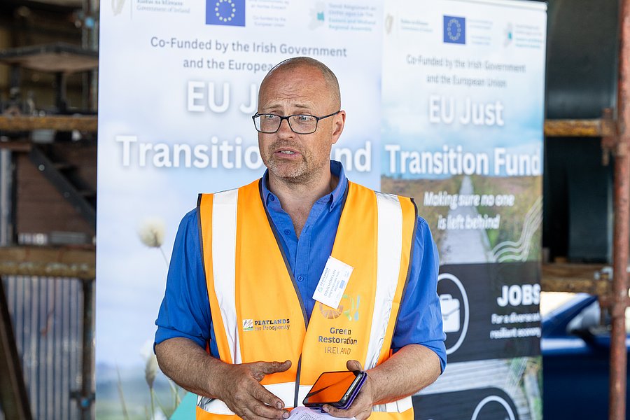 A photo of Dr. Doug McMillan at the launch of the Peatlands for Prosperity event funded by the EU Just Transition Programme. It shows a middle-aged man with a shaved head and blue eyes. He wears glasses, a blue shirt and an orange safety west. He stands in front of a roll-up of the EU Just Transition Fund.