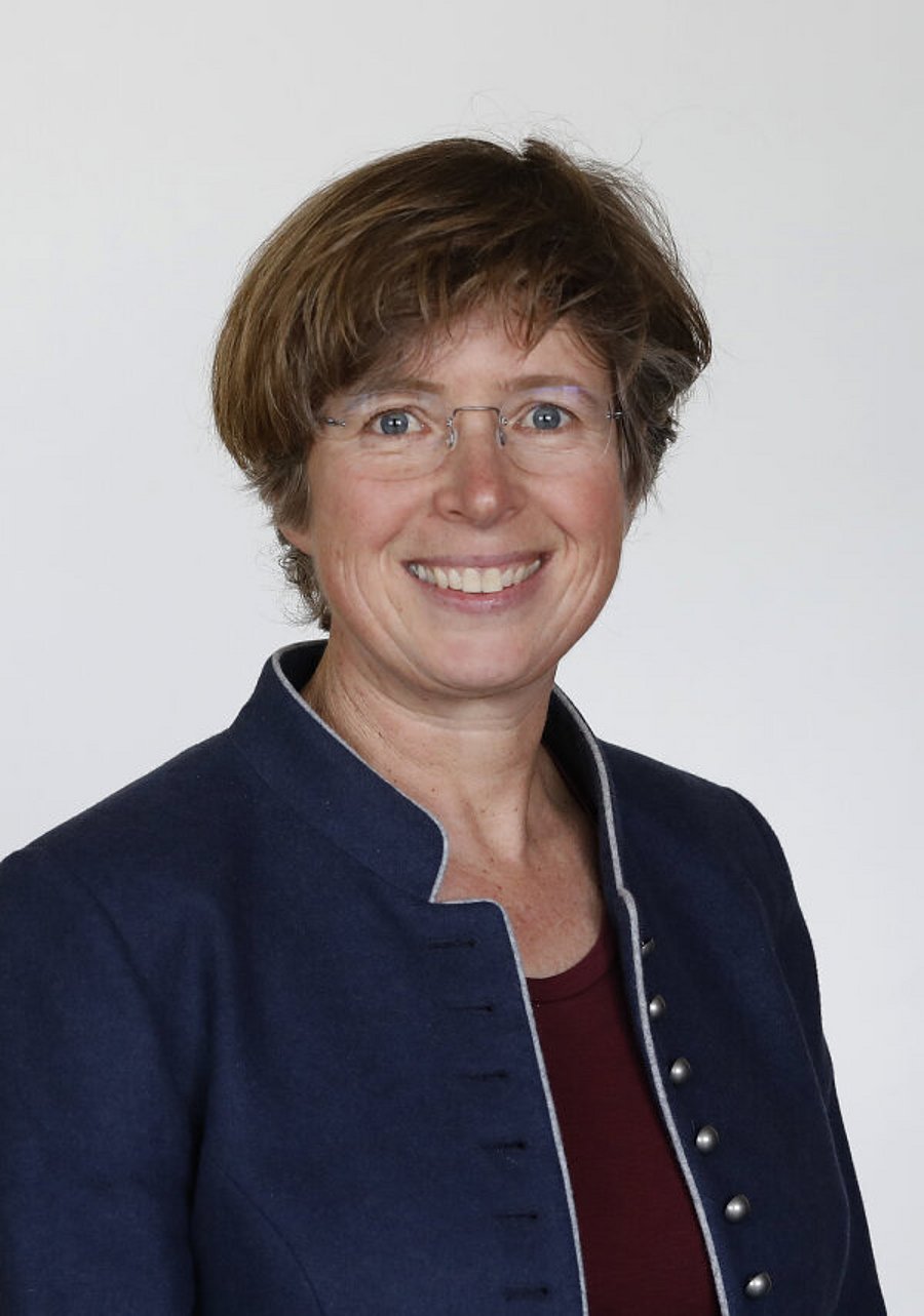 The photo shows a smiling woman with short brown hair. She is wearing rimless glasses and a dark blue blazer with a burgundy shirt underneath. 