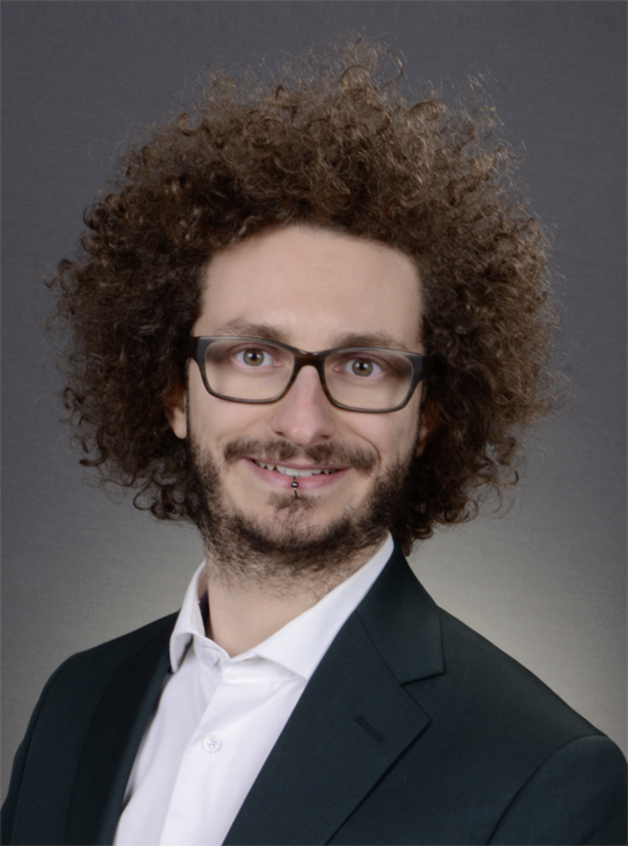 A photo of Stephen Wirt. It shows a smiling young man with brown curly hair, a full beard, brown eyes, and a piercing in his mouth. He is wearing glasses, a white shirt, and a black jacket.