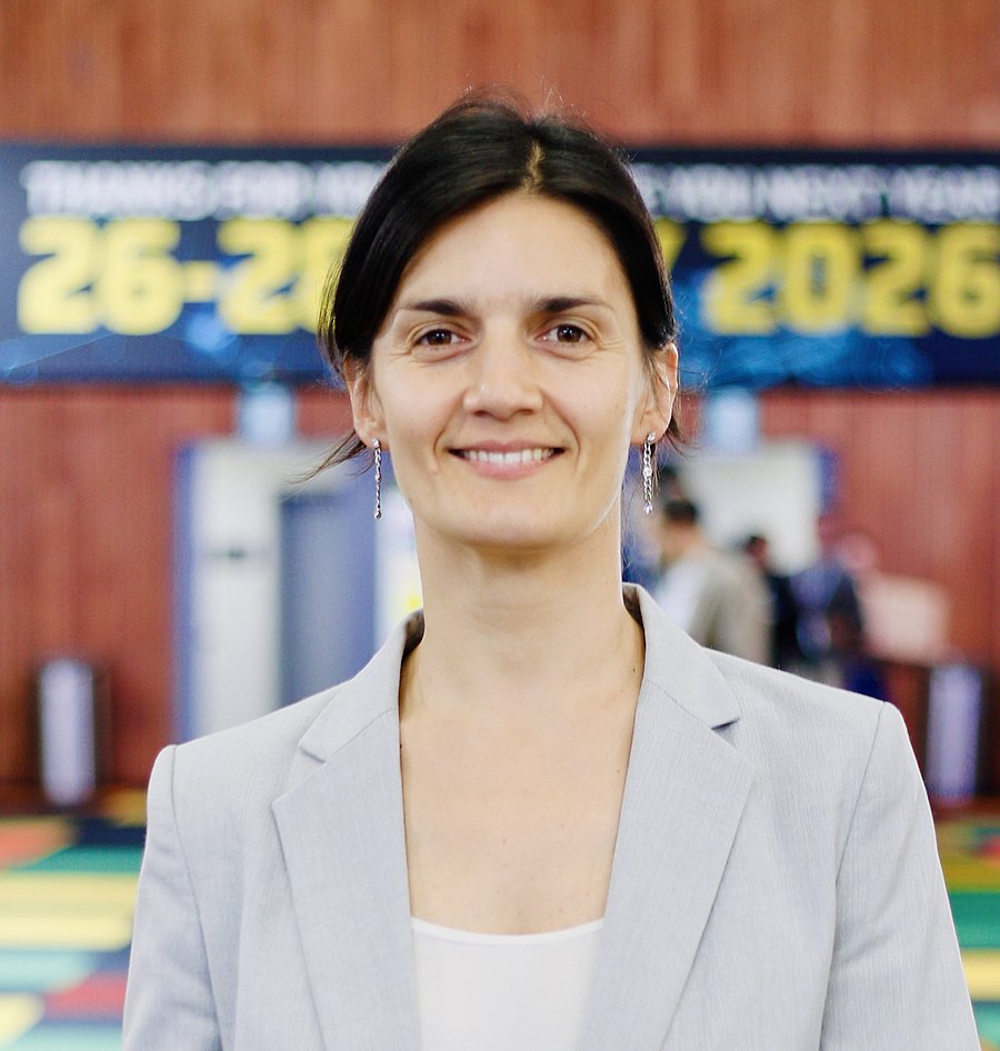 White woman with brown hair tied in a ponytail, wearing a white shirt and grey blazer, standing in front of a colourful background.