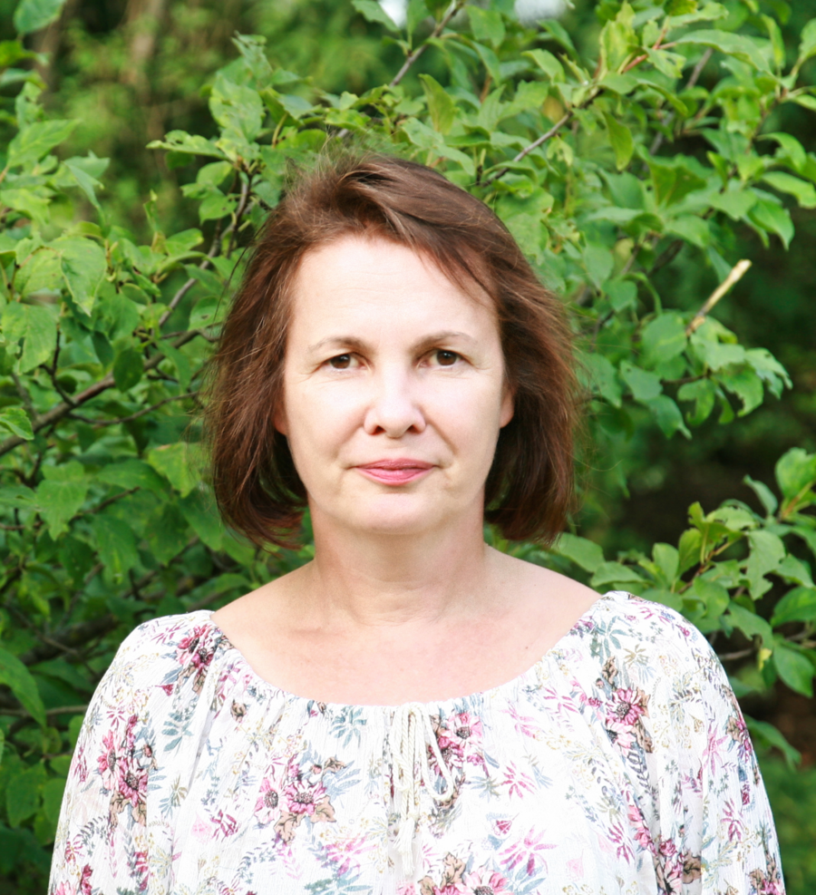 A photo of Katrin Kuka. It shows a slightly smiling woman wearing a white blouse with flowers, standing outside in front of a tree.