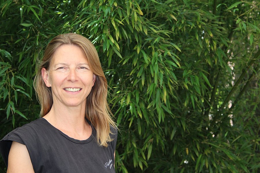 A photo of Maren Langhof. It shows a smiling woman with blonde hair. She wears a black, sleeveless shirt. She stands in front of shrub. 