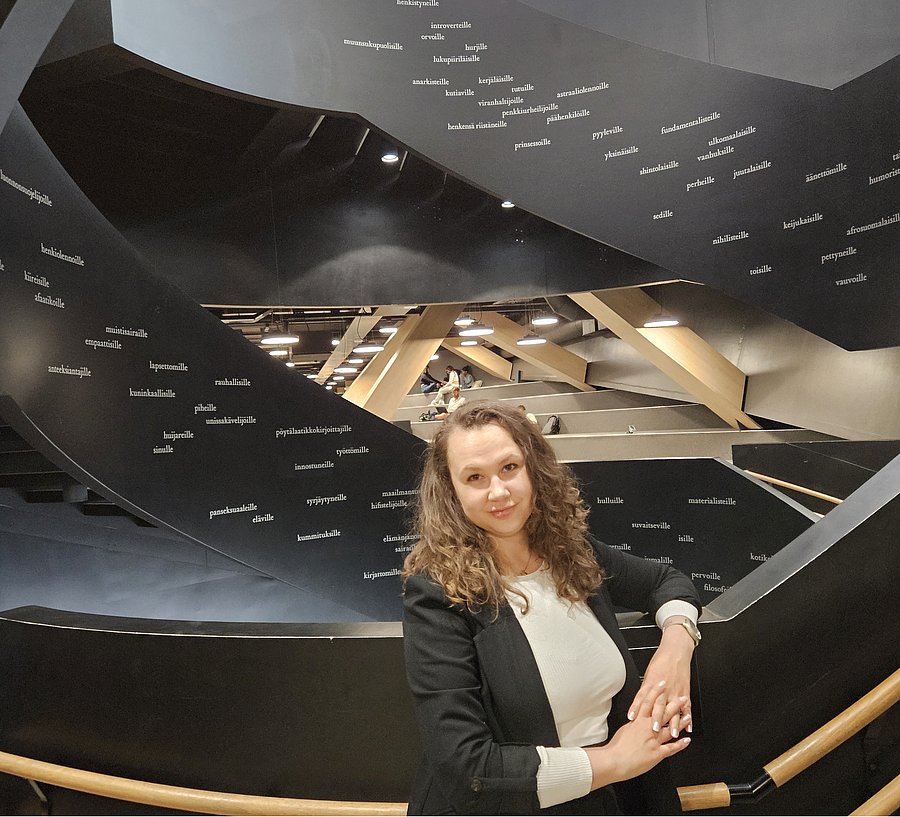 A photo of Pauline Rajewicz. It shows a woman with long brown hair standing in an auditorium. She wears a black suit over a white shirt.
