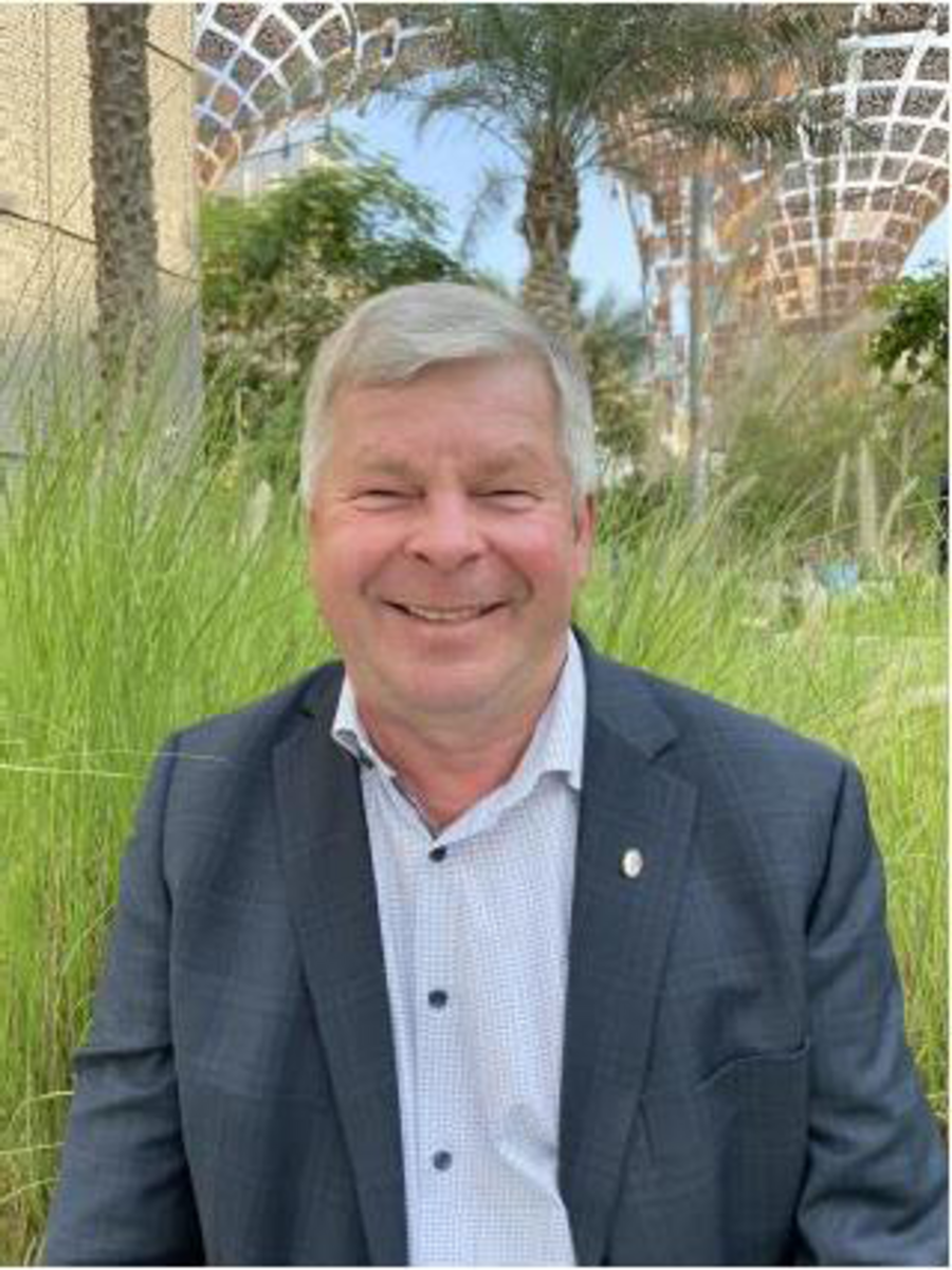 A photo of Wolfgang Zornbach. It shows a smiling older man with grey hair wearing a suit jacket over a white shirt. He stand in front of tall grass. Further away in the background a pavilion structure, a palm tree and other trees are visible.