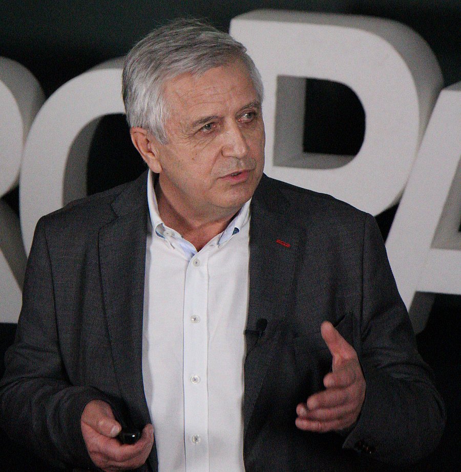 A photo of Francis Bucaille. It shows an older man with grey hair giving a presentation. He wears a black suit over a white shirt.
