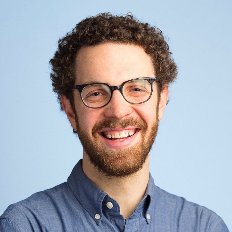 A photo of Chris Tolles. It shows a smiling man in his late thirties. He has brown, curly hair, a beard and blue eyes. He wears a blue shirt and glasses. The background of the photo is a light blue.