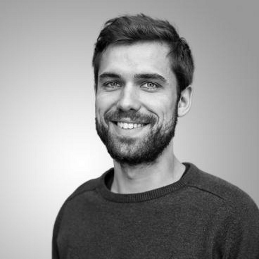 A black and white photo of Aaron Scheid. It shows a smiling man with short hair and a full beard, wearing a sweater.