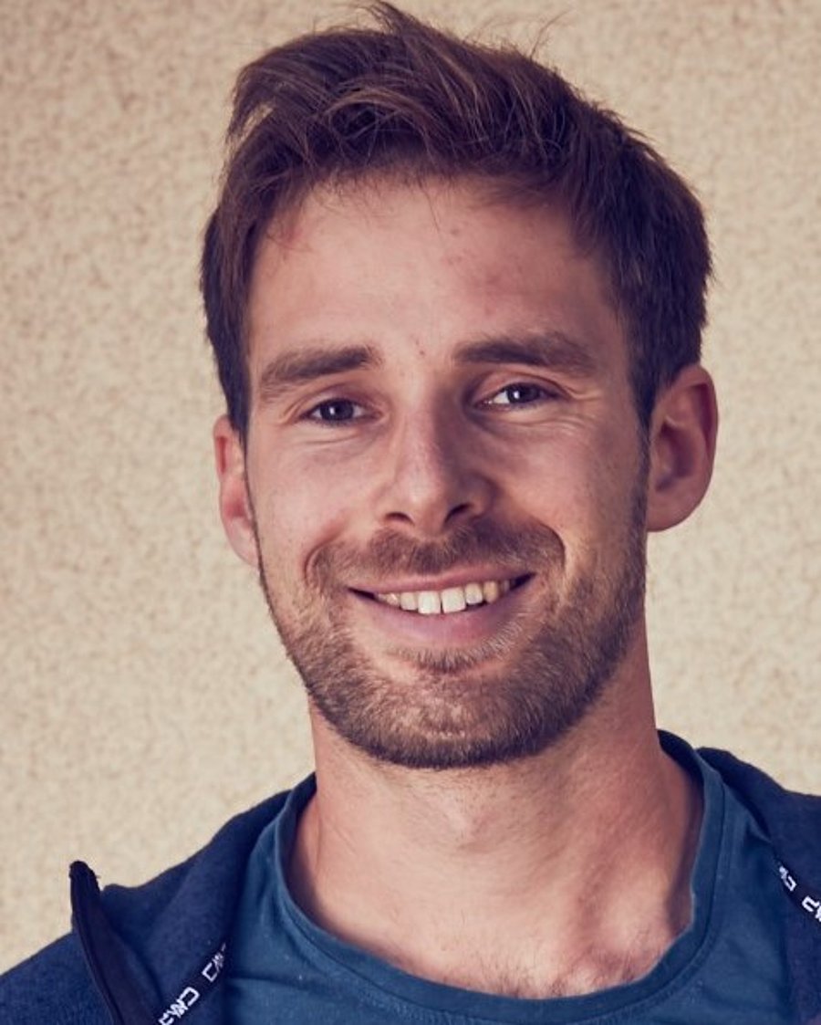 A photo of Lucas Kohl. It shows a smiling man with brown hair and a full beard, looking straight into the camera. He wears a blue jacket over a blue t-shirt. He stands in front of a beige wall.