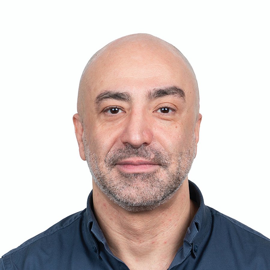 A Middle Eastern man with a shaved head, wearing a blue shirt, in front of a white background.
