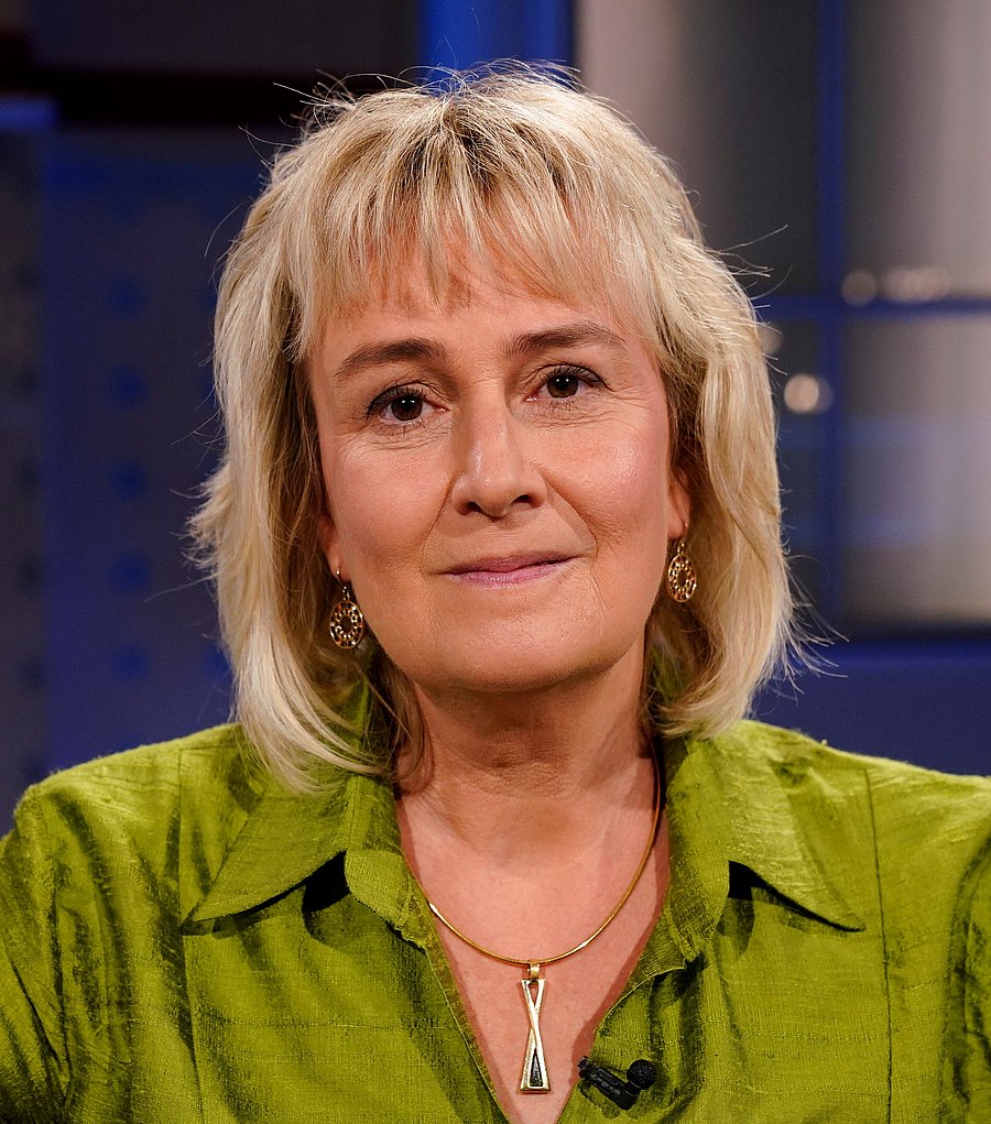 Professor Dr Claudia Kammann, who has shoulder-length blonde hair and brown eyes, looks directly into the camera. She is wearing a light green blouse, as well as earrings and a necklace with an elongated pendant. The background is blurred in shades of blue.
