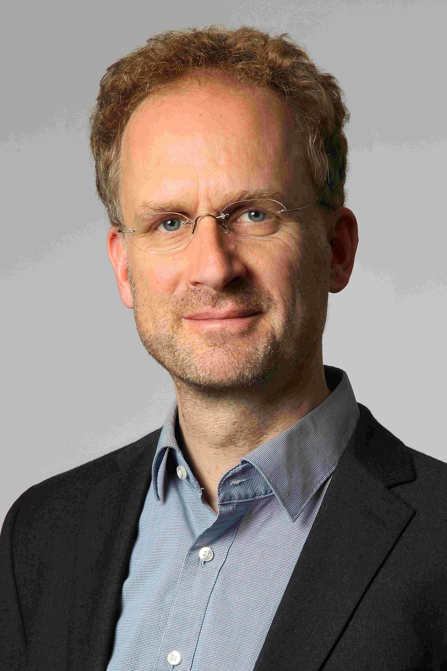 A photo of Lambert Schneider. It shows a smiling man in his forties. He has blonde, greying hair and blue eyes. He wear glasses, a black jacket and a light blue shirt.