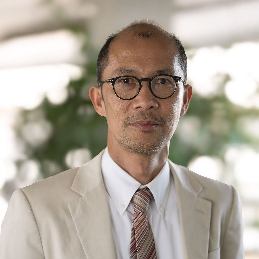 A photo of Oliver Oliveros. It shows a balding man with dark hair looking straight into the camera. He wears a light beige suit jacket over a white shirt with a tie. He also wears glasses.