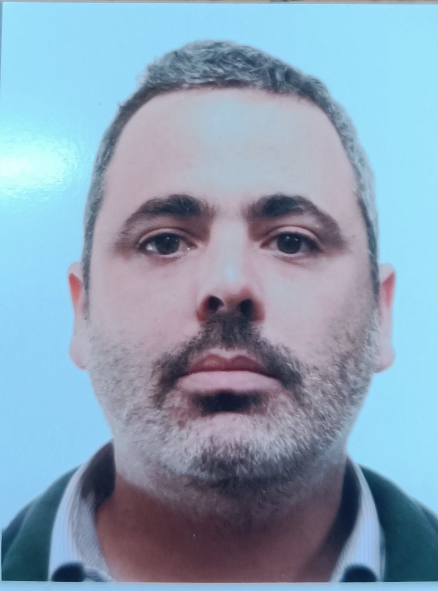 A photo of Agustin de Prado. It shows a bearded man with dark and grey hair, and dark eyes. He wears a green sweater over a striped shirt. The background of the photo is blue.