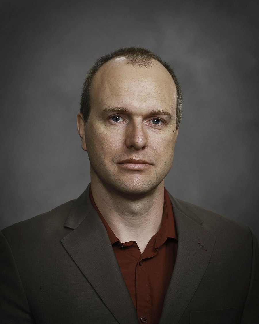 A photo of Roland Kröbel. It shows a man in his late thirties. He has dark hair and dark eyes. He wears a brown suit jacket over a dark red shirt. The background of the photo is a grey.