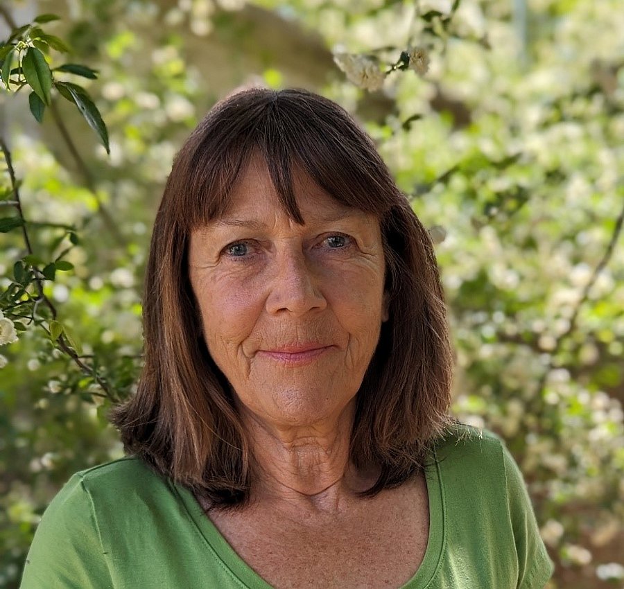 A photo of Elke Noellenmeyer. She has shoulder-length brown hair and is wearing a green shirt. She is smiling and standing in front of a tree.