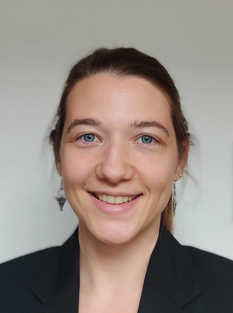 A photo of Ramona Stossberger. It shows a smiling young woman with dark blond hair and blue eyes. She wears a black jacket and earrings. She stands in front of a white background.