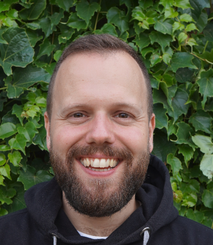 A photo of Julian Kremers. It shows a smiling man with brown hair and a full beard looking straight into the camera. He wears a black hoodie and stand in front of a hedge.
