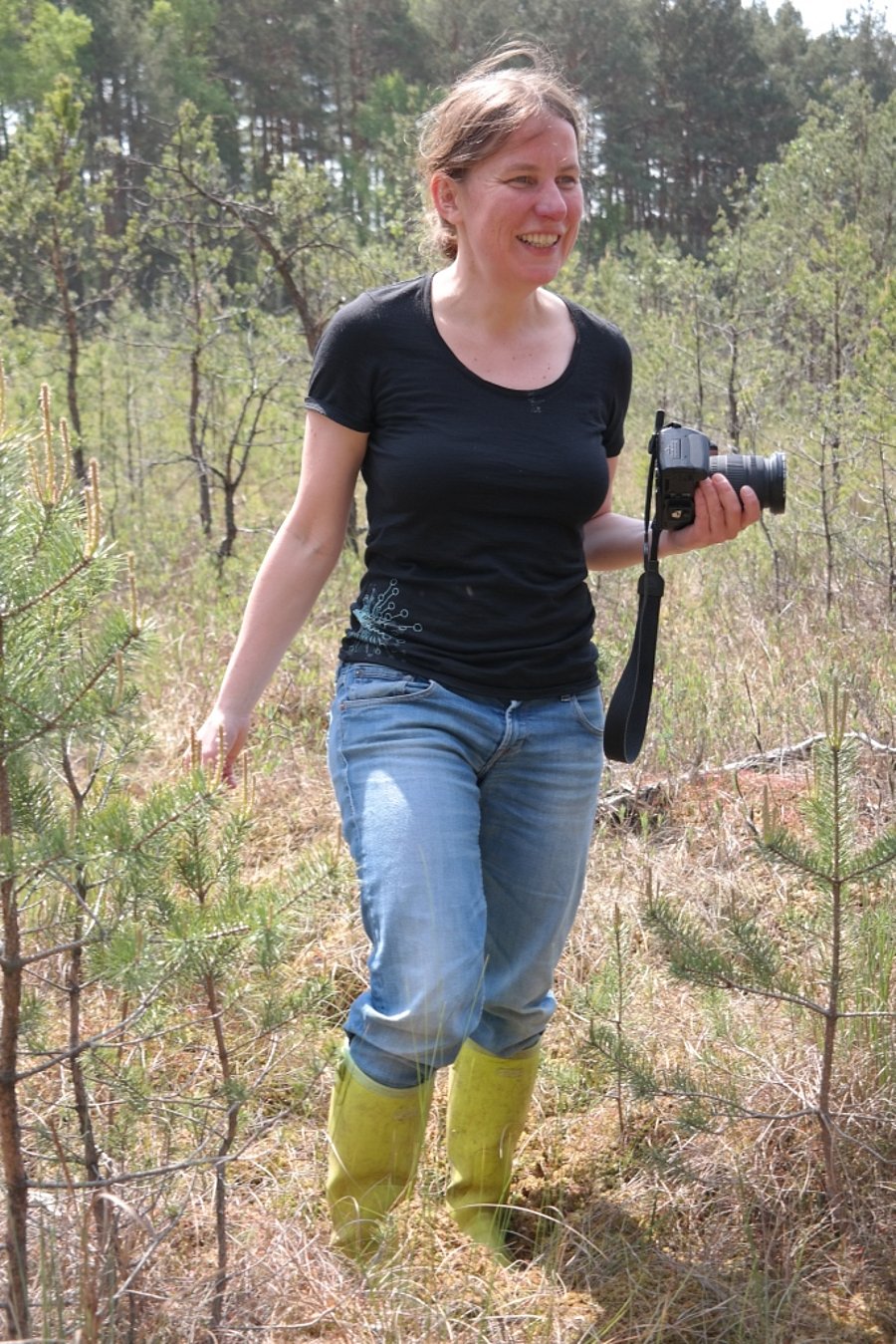A photo of Bärbel Tiemeyer. It shows a woman with brown hair wearing blue jeans, a black t-shirt and rubber boots, holding a camera in her left hand. She is standing in a clearing surrounded by young trees. A forest is visible in the background.