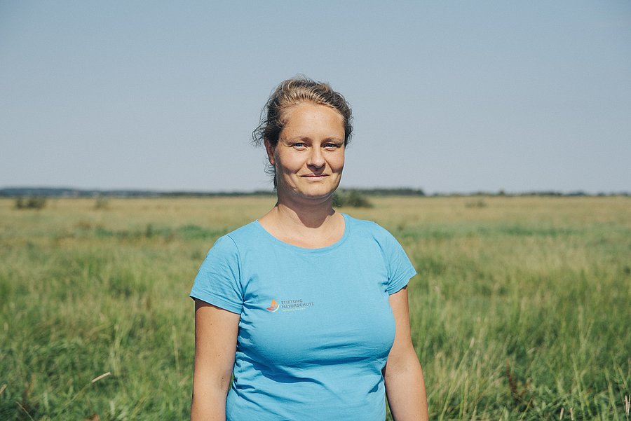 A photo of Wiebke Schuster. It shows a white woman with blonde hair tied into a ponytail, wearing a blue t-shirt in front of a nature background.